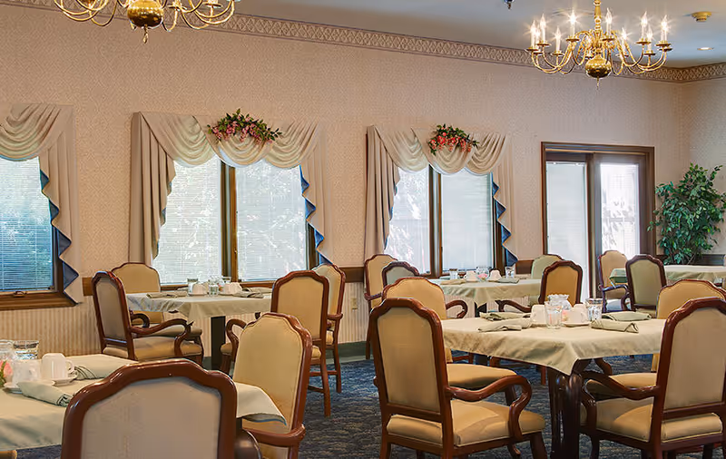 A dining room with several tables covered with beige tablecloths and set with cups, glasses, and napkins. The room has upholstered wooden chairs around the tables, large windows with decorative curtains, a blue carpeted floor, and two chandeliers hanging from the ceiling. There is a potted plant in the corner near a glass door.