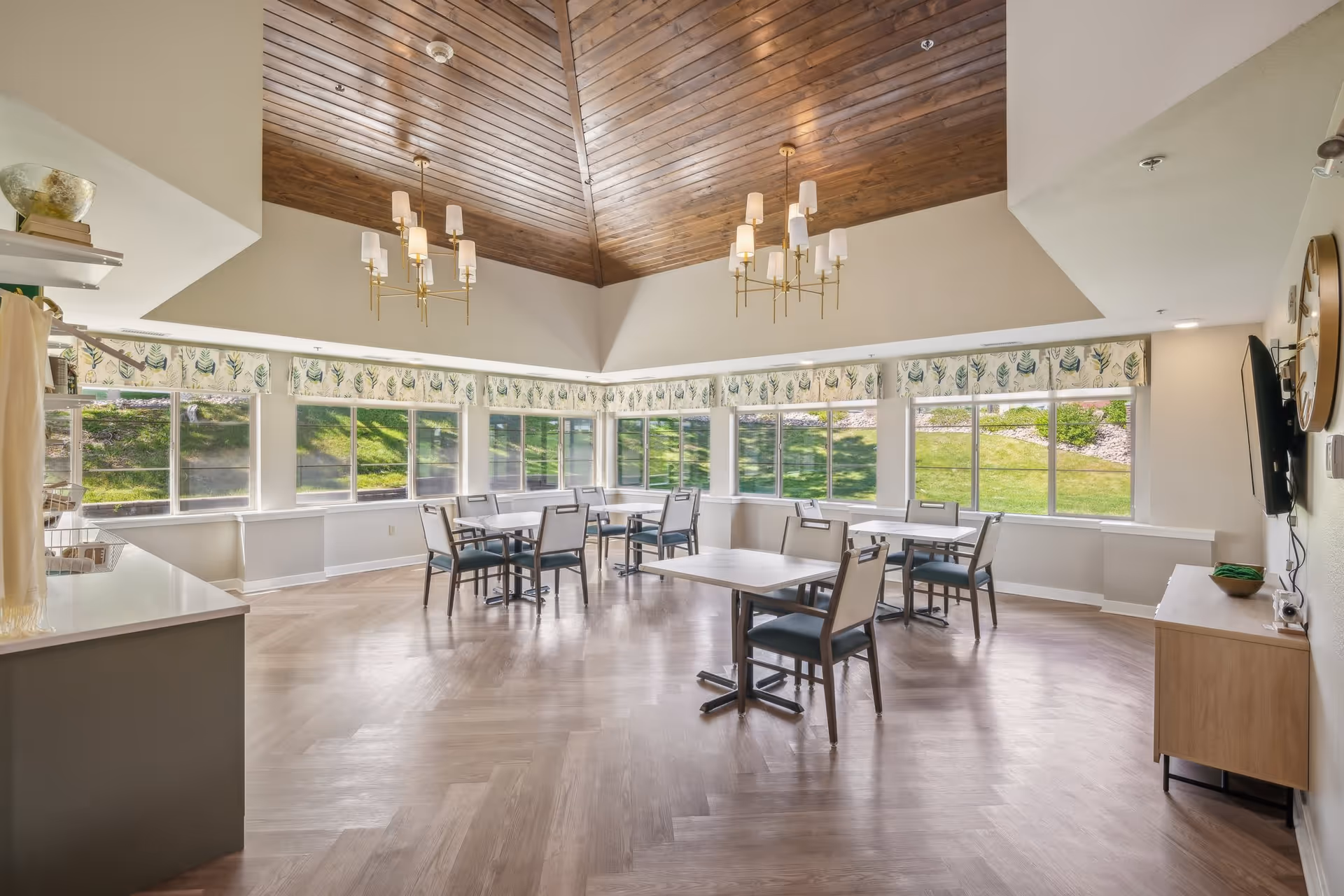 A bright dining room with multiple square tables and chairs arranged neatly. The room features large windows with leafy patterned valances, allowing natural light to fill the space and providing a view of green grass outside. The ceiling is vaulted with wooden paneling and two modern chandeliers hanging down. There is a TV mounted on the right wall above a wooden cabinet, and a counter with shelves on the left side.
