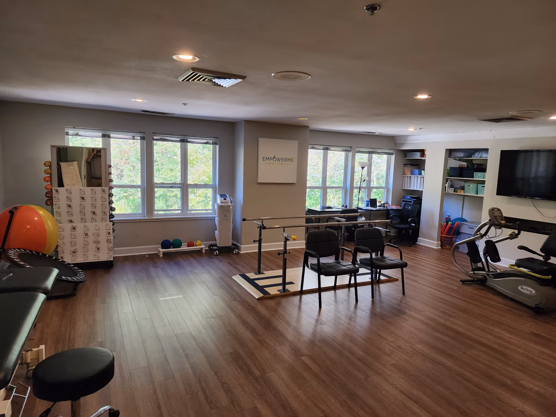 A spacious wellness or physical therapy room with wooden flooring, large windows letting in natural light, exercise equipment including a stationary bike, therapy balls, parallel bars for walking exercises, chairs, and shelves with various supplies. A sign on the wall reads 'EmpowerMe Wellness'.