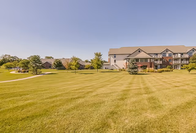 A large, well-maintained grassy lawn with a few small trees scattered throughout. In the background, there is a multi-story building with balconies and a smaller building partially visible to the left. The sky is clear and blue.