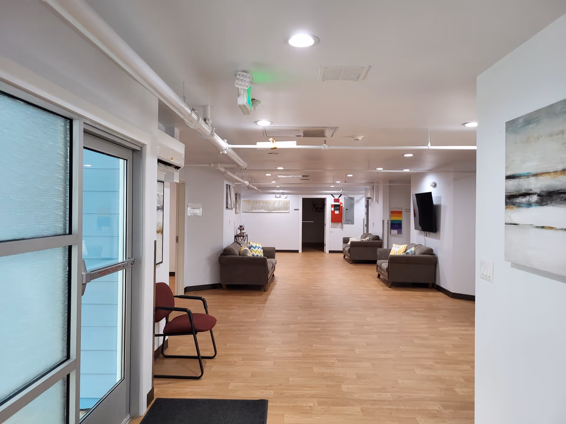 Interior hallway of a senior living facility with light wood flooring, white walls, and ceiling lights. The hallway features several brown sofas with colorful pillows along the walls, a mounted flat-screen TV, and abstract artwork. A glass door with frosted panels is visible on the left side, and a maroon chair is placed near the door.