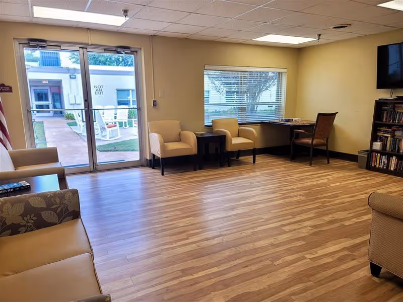 A well-lit room with wooden flooring featuring several armchairs, a small table, a desk with a chair, a bookshelf filled with books, and a wall-mounted television. There is a large window with blinds and glass doors leading outside to a patio area with white rocking chairs.