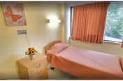 Simple single-occupancy bedroom with a single bed covered in a pink bedspread, a nightstand with a vase of flowers, wall lamp, artwork, and a window with pink curtains.