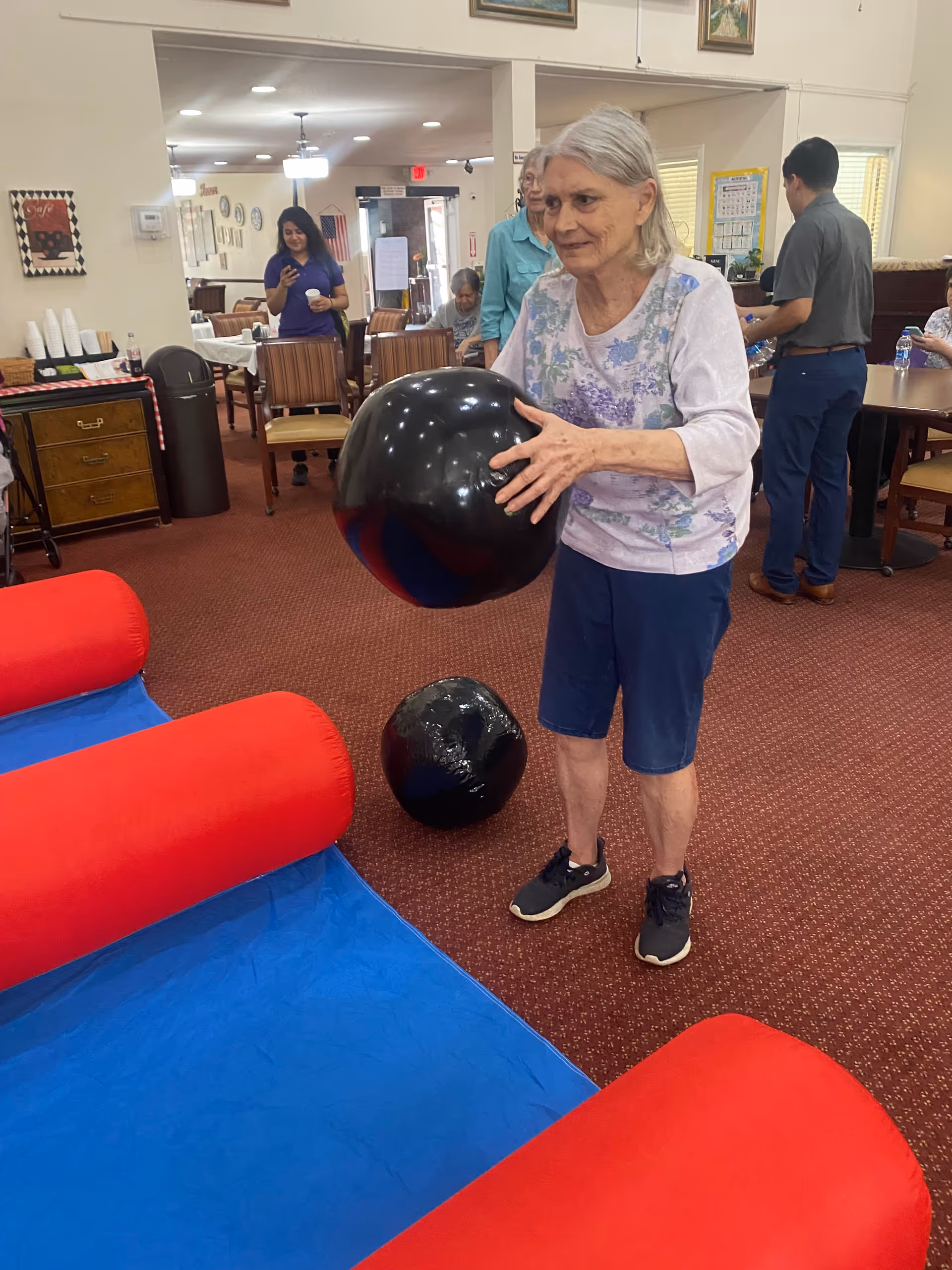 An elderly woman holding a large black ball indoors, standing on a carpeted floor near a blue and red inflatable structure. Several other people are in the background, some seated and some standing, in a room with tables and chairs.