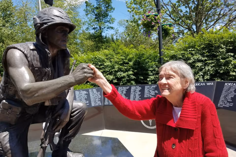 An elderly woman wearing a red sweater is smiling and holding hands with a bronze statue of a soldier kneeling. The background includes a memorial wall with names and greenery under a clear blue sky.