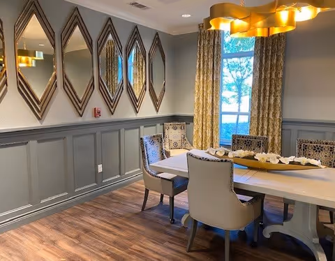 A dining room with a rectangular white table surrounded by six upholstered chairs. The room features wood flooring, a set of diamond-shaped mirrors on one wall, patterned curtains framing a window, and a modern gold chandelier hanging above the table. A decorative centerpiece is placed on the table.