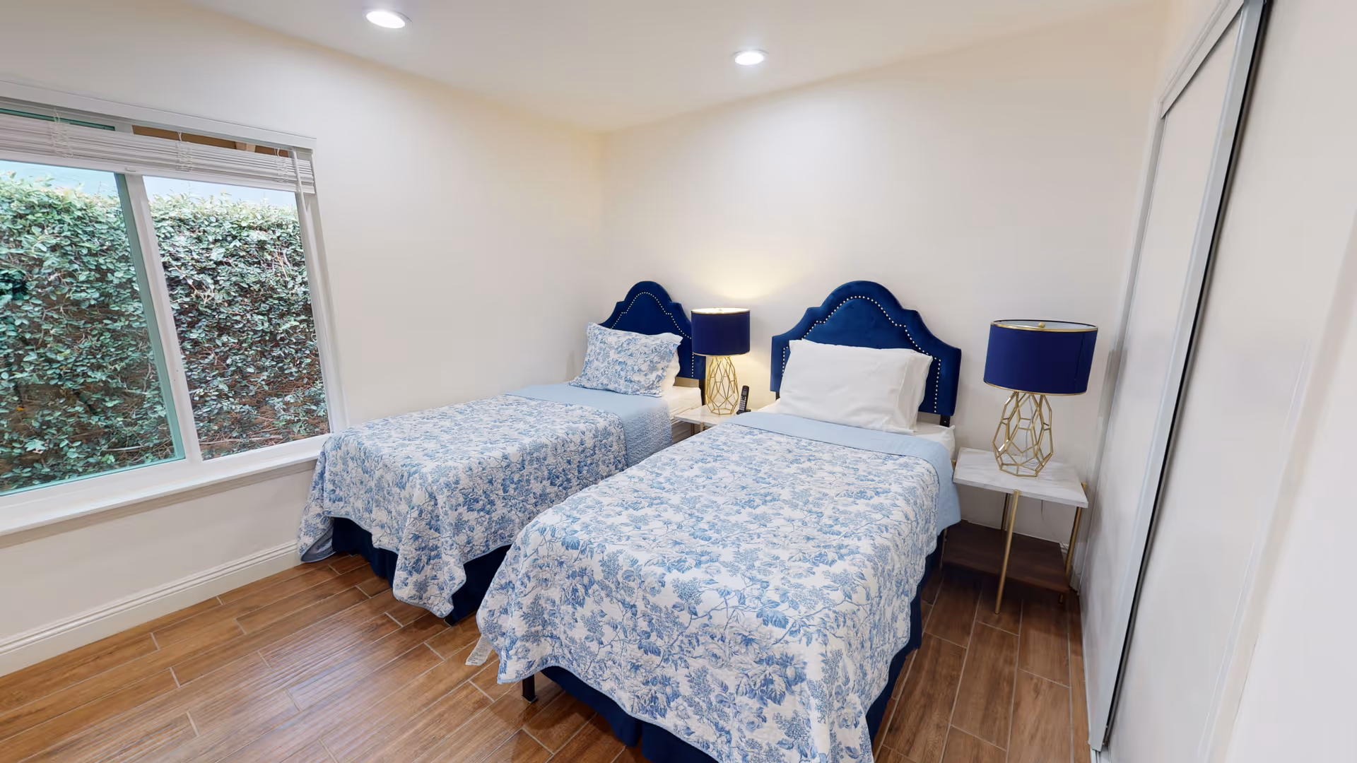 A bedroom with two twin beds featuring blue and white floral bedspreads and blue upholstered headboards. Each bed has a white pillow and a blue lamp with a gold geometric base on a white nightstand between them. There is a large window with a view of greenery outside and wooden flooring.