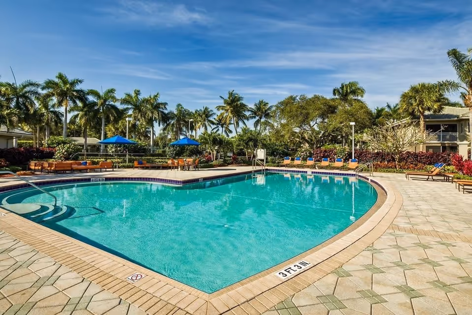 Outdoor swimming pool with clear blue water surrounded by a tiled deck. There are lounge chairs and tables with blue umbrellas around the pool. Palm trees and other greenery are visible in the background under a blue sky with some clouds.