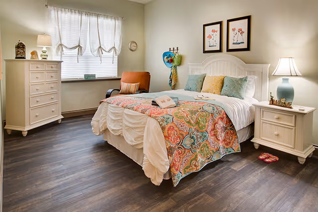 Bright furnished bedroom with a bed topped by a colorful quilt, two nightstands with lamps, a dresser, an armchair, and wood-look flooring.