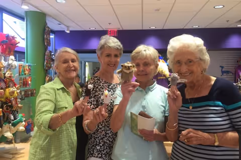 Four elderly women standing inside a colorful store or gift shop, each holding a small puppet or figurine and smiling at the camera. The background shows shelves with various items and a purple wall.
