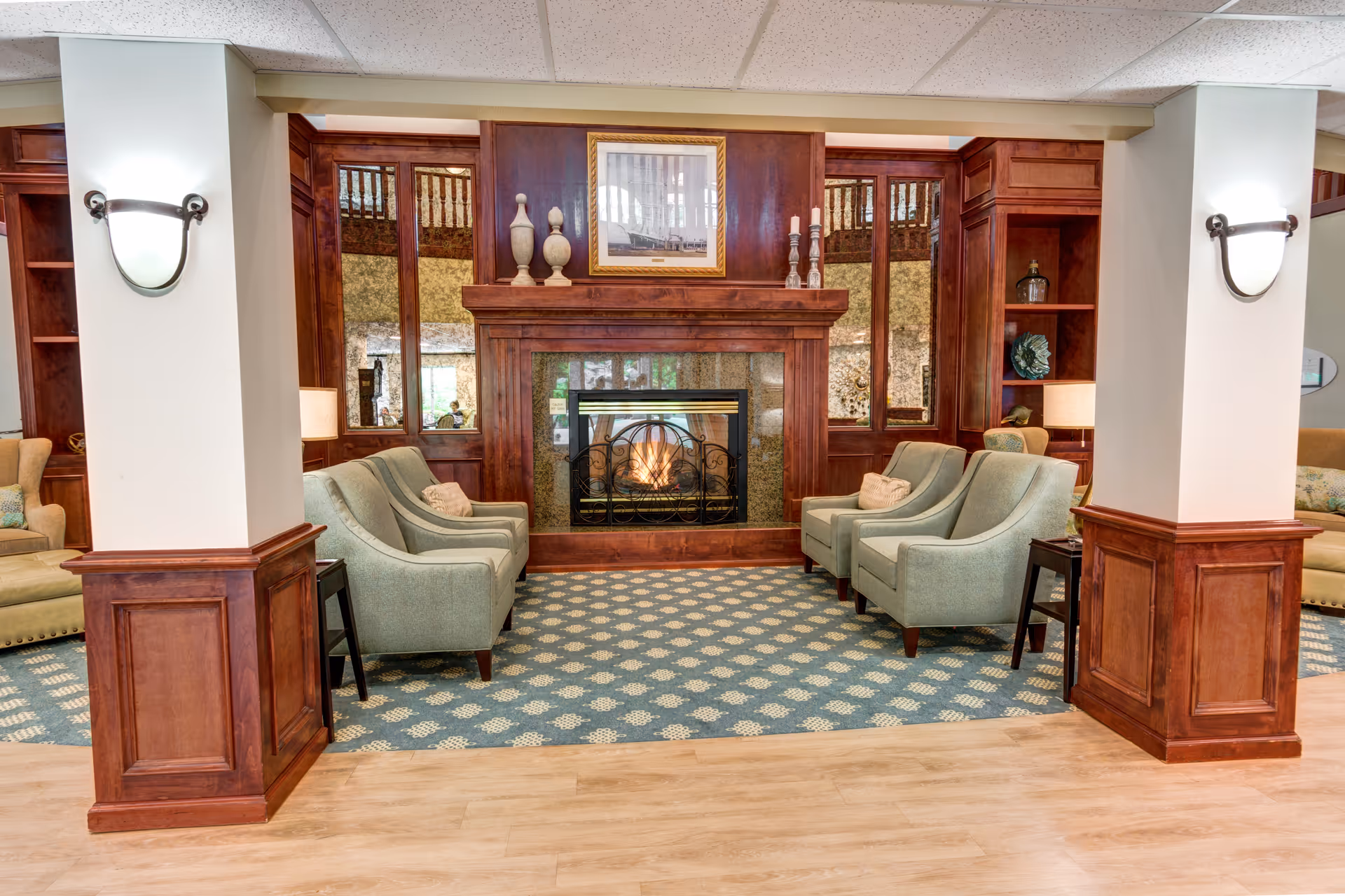 A cozy senior living facility lounge area with a central fireplace surrounded by wooden paneling and mirrors. The room features four light green armchairs arranged in two pairs facing each other on a patterned blue carpet. There are two white pillars with wall-mounted lights, and additional seating with beige sofas is visible on the sides.
