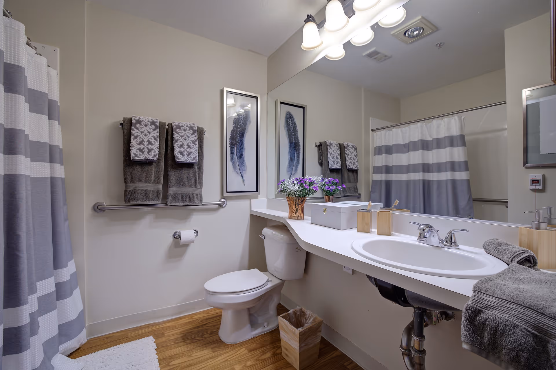 A clean and modern bathroom with a toilet, a white sink with a countertop, and a large mirror above it. There are two framed feather artworks on the wall, a towel rack with two patterned towels, a striped shower curtain, a small trash bin, and a vase with purple flowers on the countertop.