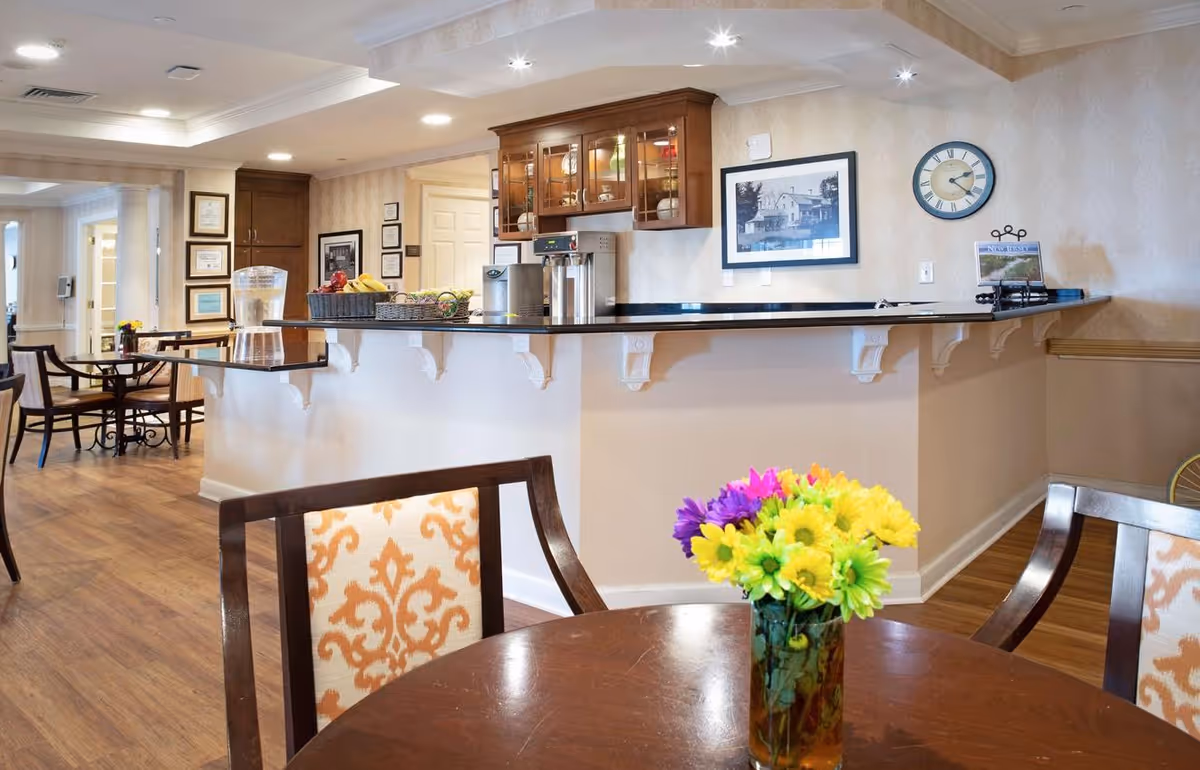 Interior view of a senior living facility dining area with wooden tables and chairs. A round table in the foreground has a vase with colorful flowers. In the background, there is a counter with a black countertop, a water dispenser, baskets of fruit, and wooden cabinets mounted on the wall. The walls are decorated with framed pictures and a clock.