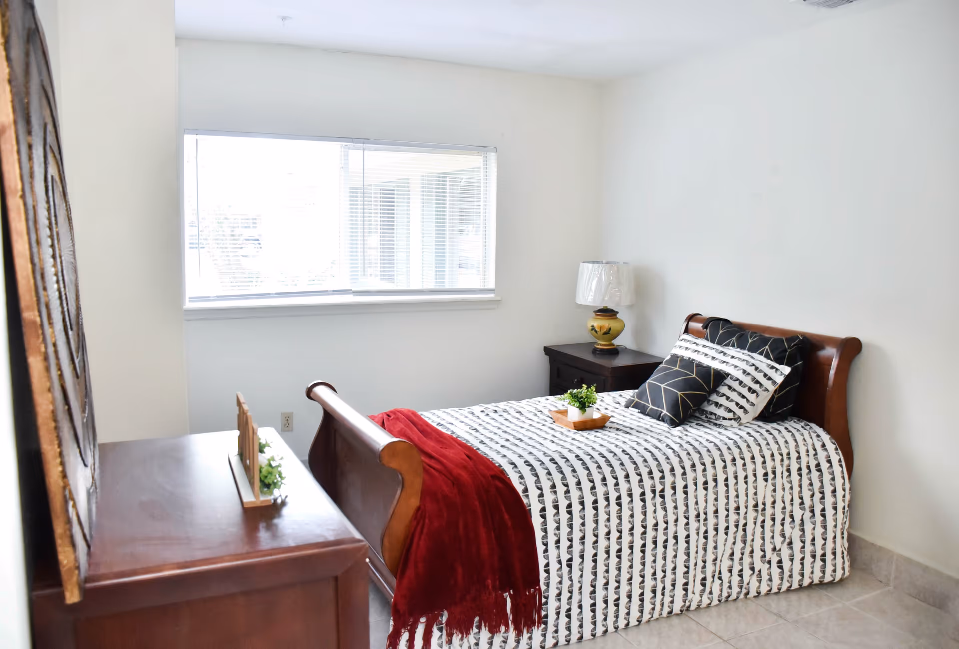 A bright bedroom with a wooden sleigh bed covered in a white and black patterned bedspread, accented with a red throw blanket and decorative pillows. There is a wooden nightstand with a lamp and a small plant on it, and a wooden dresser with a decorative item on top. A large window with blinds lets in natural light.