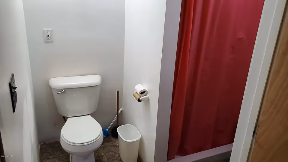 A small bathroom featuring a white toilet, a toilet paper holder with a roll of toilet paper, a white trash bin, a toilet brush, and a red shower curtain partially visible on the right side.