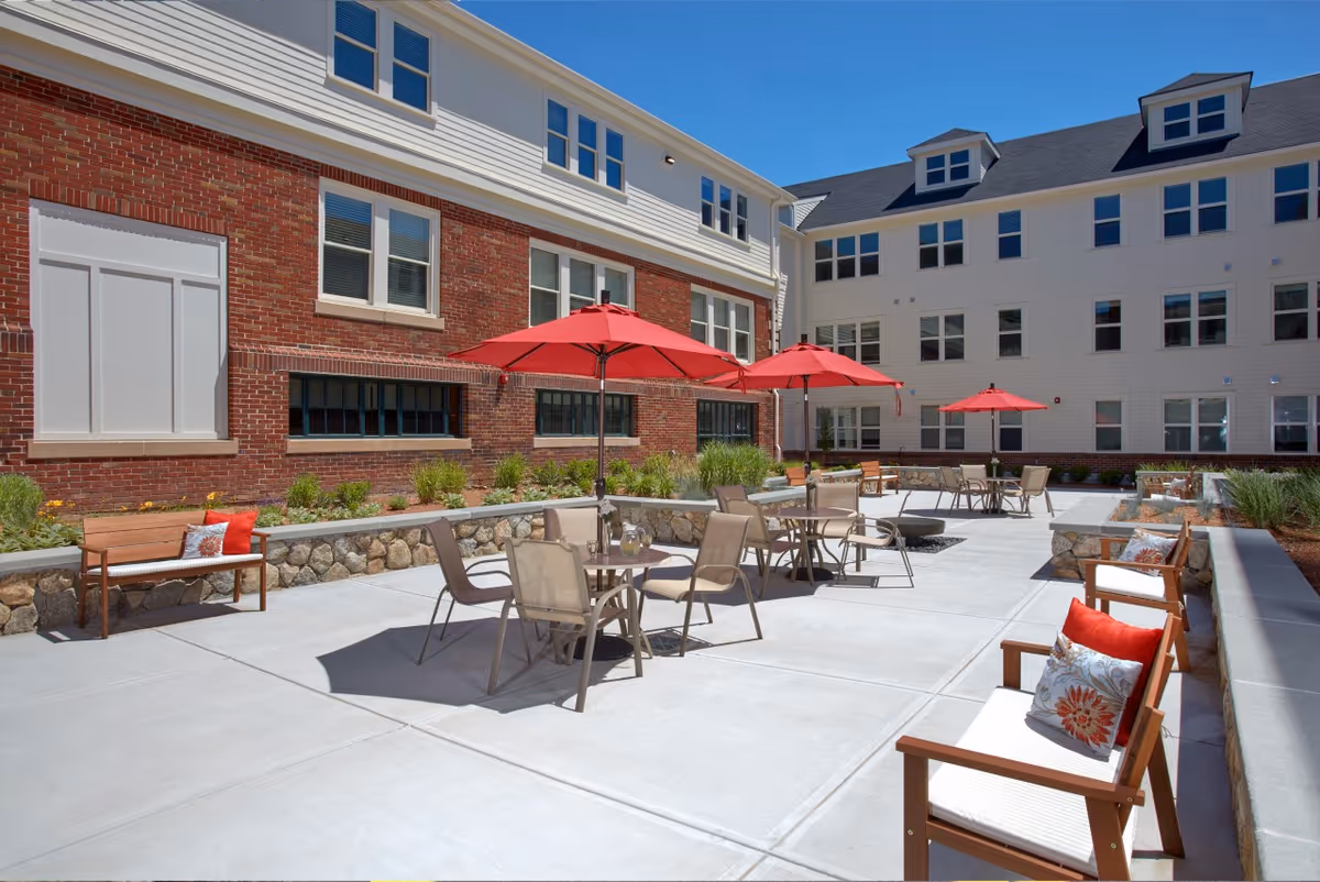 Outdoor patio area at Forestdale Park Senior Living with tables and chairs under red umbrellas, wooden benches with cushions, surrounded by a brick and white building under a clear blue sky.
