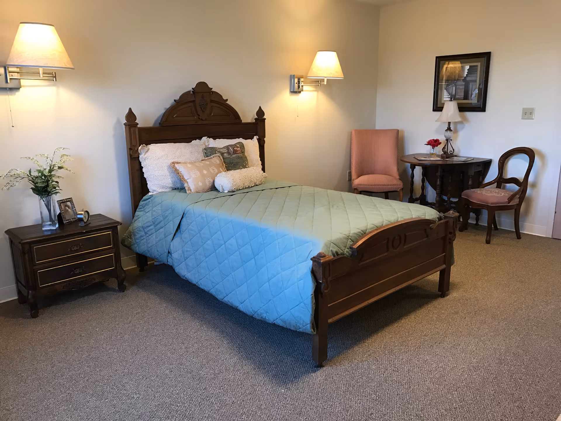 A bedroom with a wooden bed frame and a light blue quilted bedspread, decorated with multiple pillows. On the left side of the bed is a wooden nightstand with a vase of flowers and a framed photo. Two wall-mounted lamps provide lighting above the bed. On the right side of the room, there is a small round wooden table with a lamp, a vase with red flowers, and two chairs, one upholstered in pink and the other wooden with a cushioned seat. A framed picture hangs on the wall above the table.
