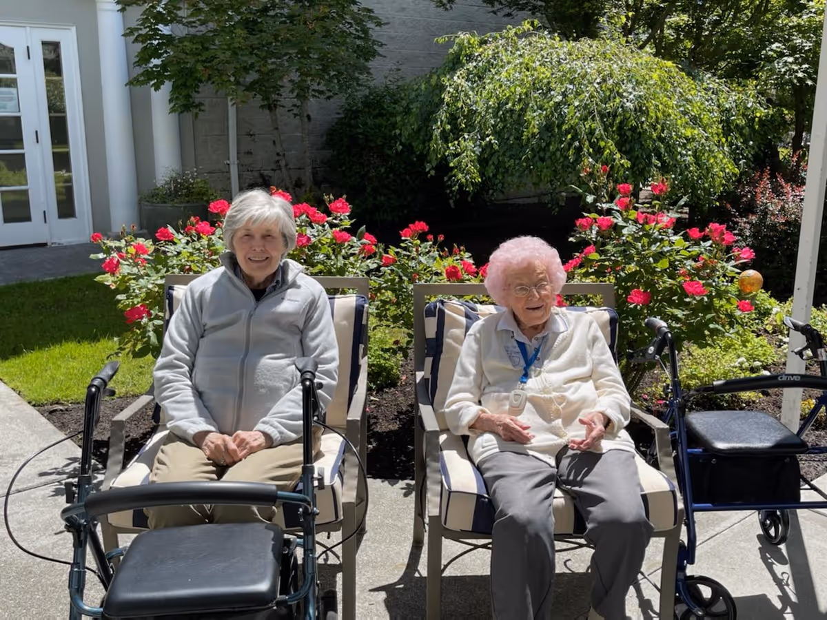 Two elderly women sitting outdoors on cushioned chairs with walkers in front of them. They are surrounded by green plants and blooming red flowers, with a building entrance visible in the background.
