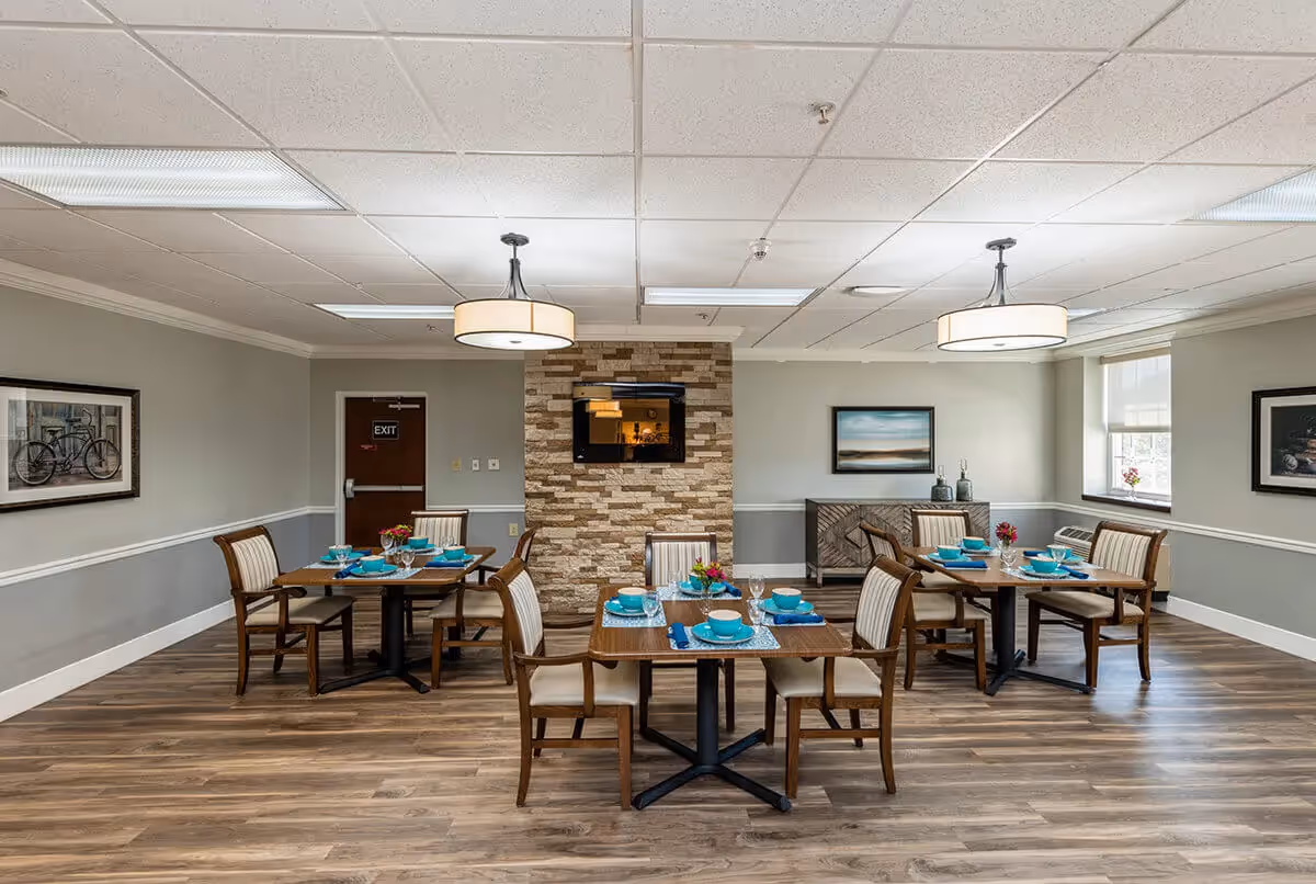 A dining room with three wooden tables, each set with turquoise dishes, glasses, and napkins. Each table is surrounded by four wooden chairs with cushioned seats and backs. The room has wood flooring, light gray walls with white trim, and a stone accent wall in the center with a mounted flat-screen TV. Two modern ceiling lights hang from a white tiled ceiling. There are framed pictures on the walls and a window letting in natural light on the right side.