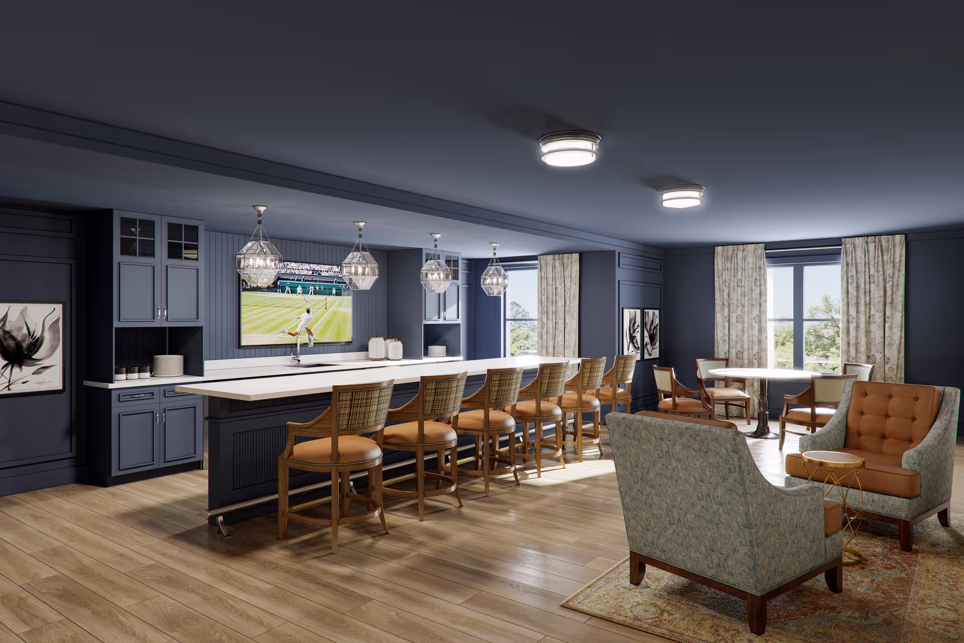 A spacious and modern common area with a long bar counter featuring eight wooden bar stools with cushioned seats. Above the counter are four geometric pendant lights. A large flat-screen TV mounted on the wall displays a tennis match. The room has dark blue walls, two windows with floral curtains, and a seating area with two upholstered armchairs and a small round table on a patterned rug. There is also a round dining table with four chairs near the windows.