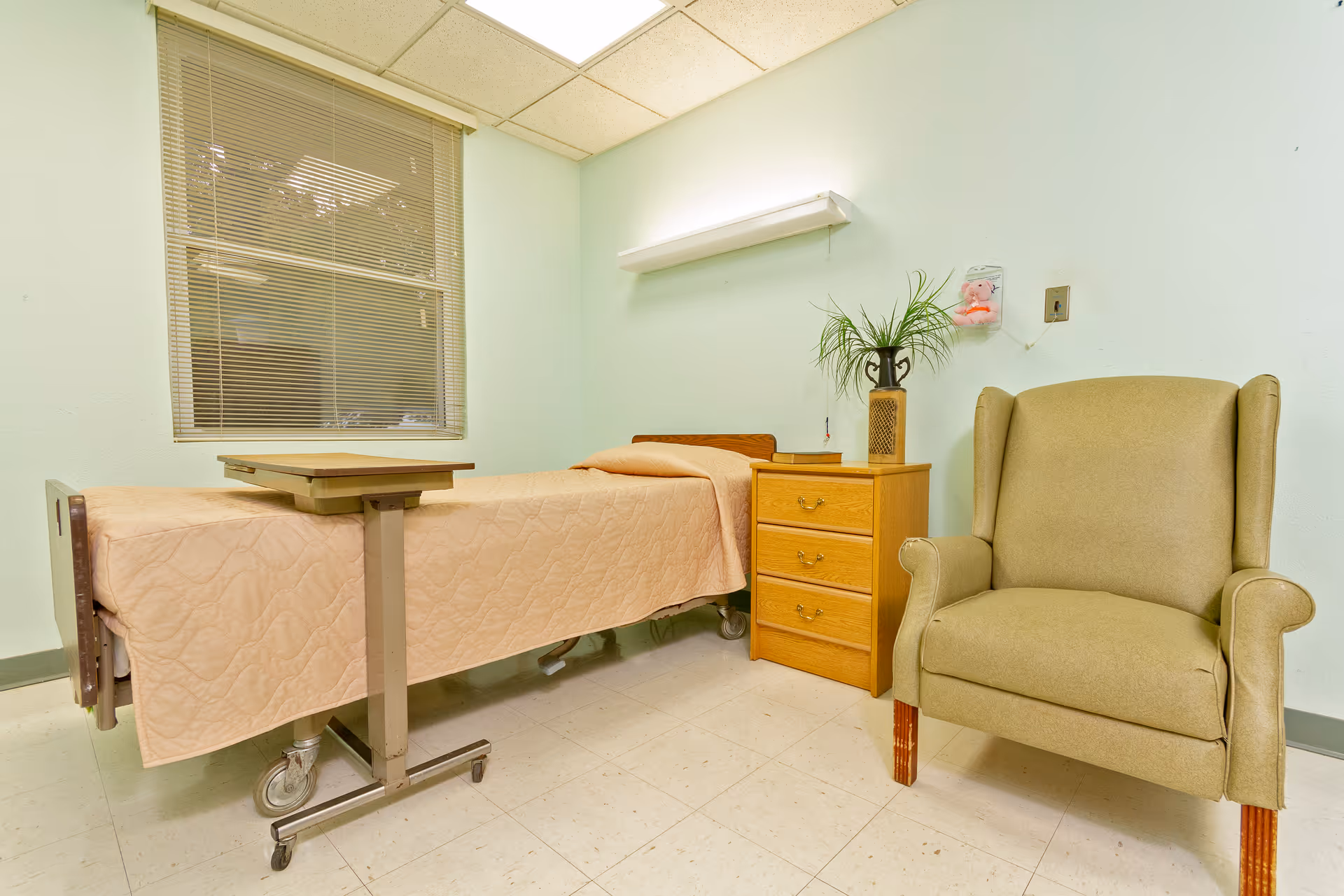 A simple nursing facility room with a single hospital bed, overbed table, wooden nightstand with a plant, and an upholstered armchair.