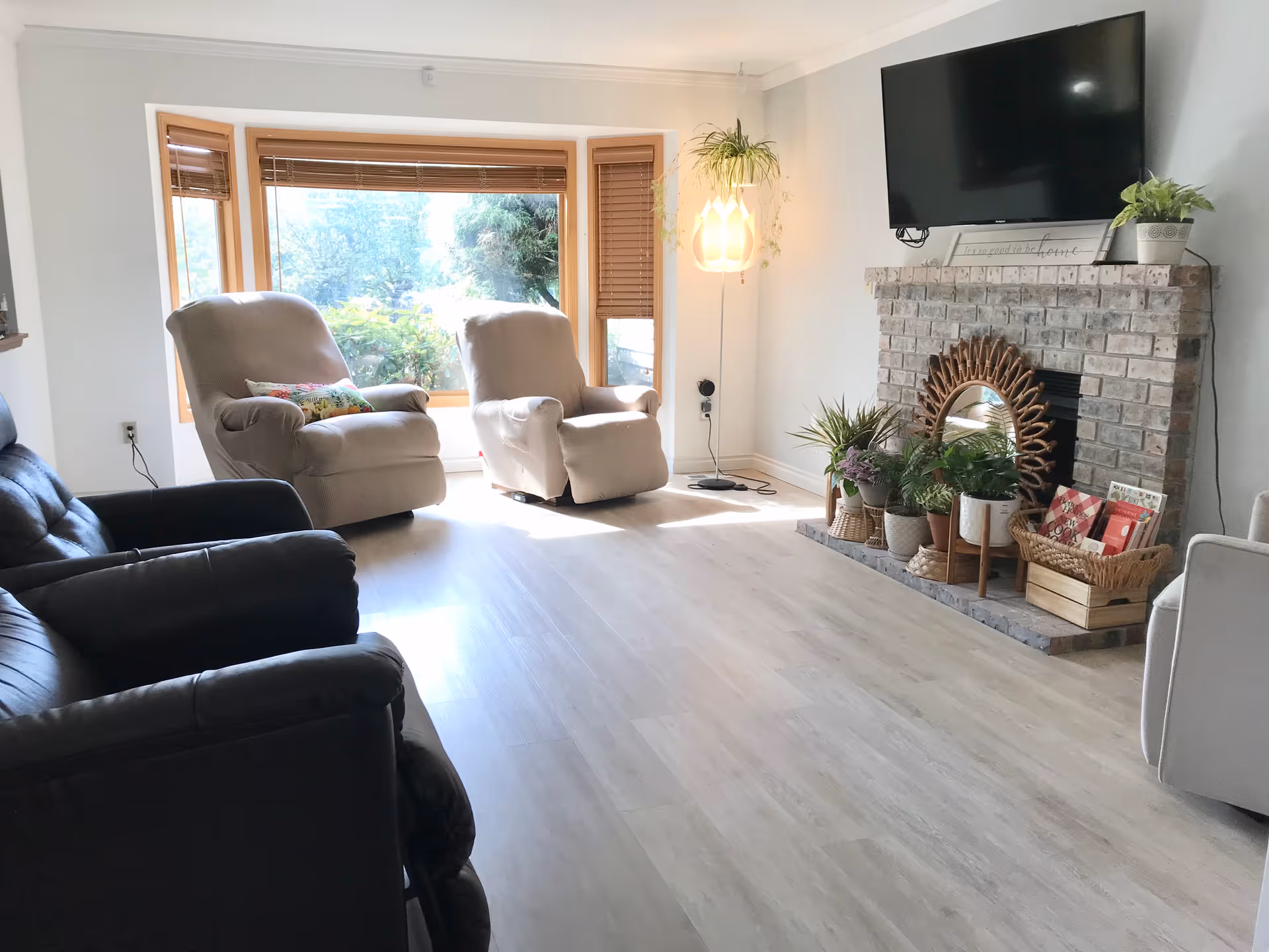 A bright living room with large windows letting in natural light, two beige recliner chairs with colorful pillows, a black leather sofa, a brick fireplace decorated with various potted plants and a decorative mirror, a flat-screen TV mounted above the fireplace, and a hanging lamp with a plant beside it.