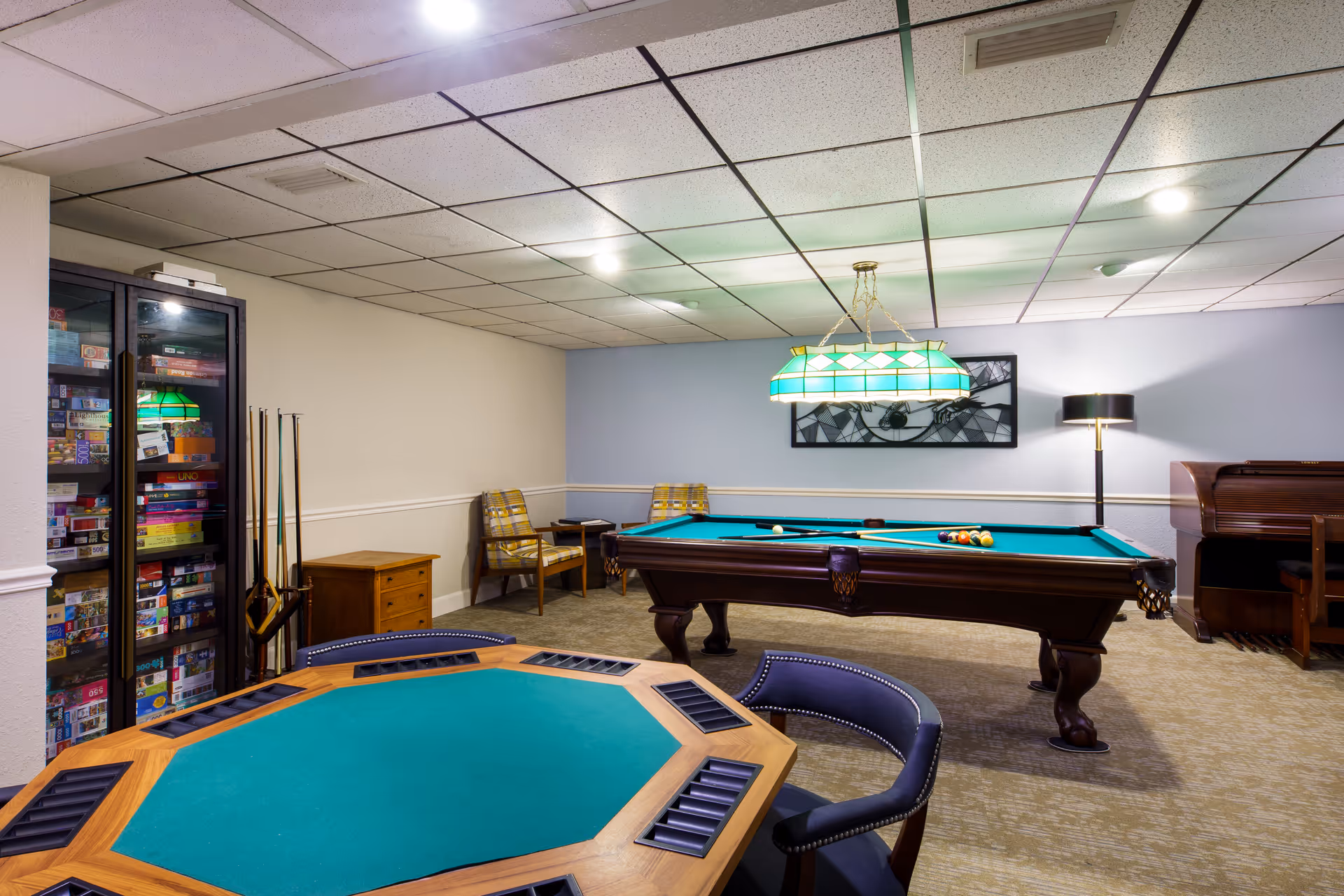 A recreational room with a pool table, a green felt card table with chairs, a cabinet filled with board games, two yellow plaid chairs with a small table between them, a floor lamp, and a piano against the wall. The room has a drop ceiling with fluorescent lighting and a green stained glass hanging light above the pool table.