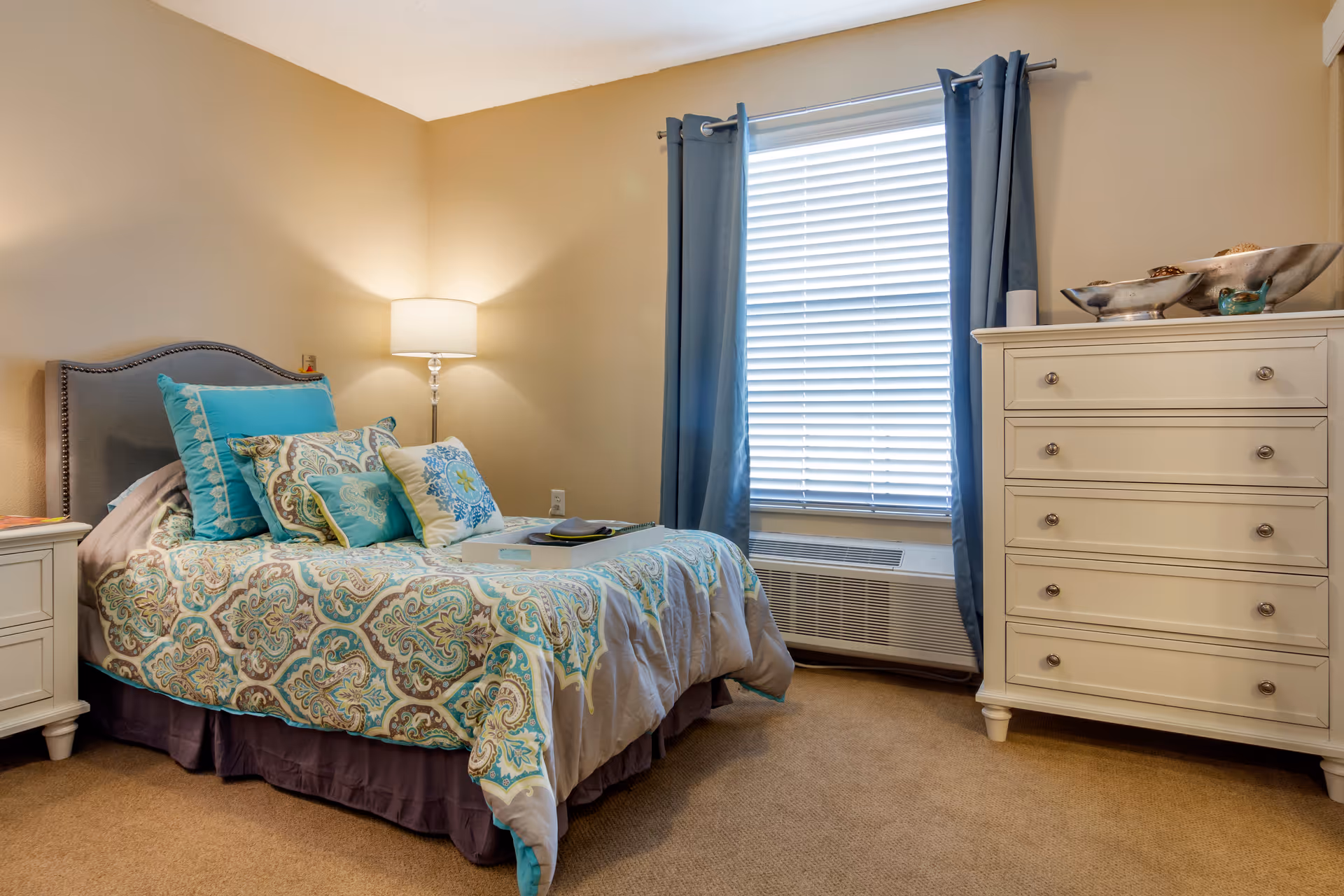 A cozy bedroom with a bed featuring a patterned comforter and multiple decorative pillows in shades of blue and beige. There is a nightstand with a lamp on the left side of the bed, a window with blue curtains and blinds, and a white dresser with decorative bowls on top on the right side of the room. The walls are painted beige and the floor is carpeted.