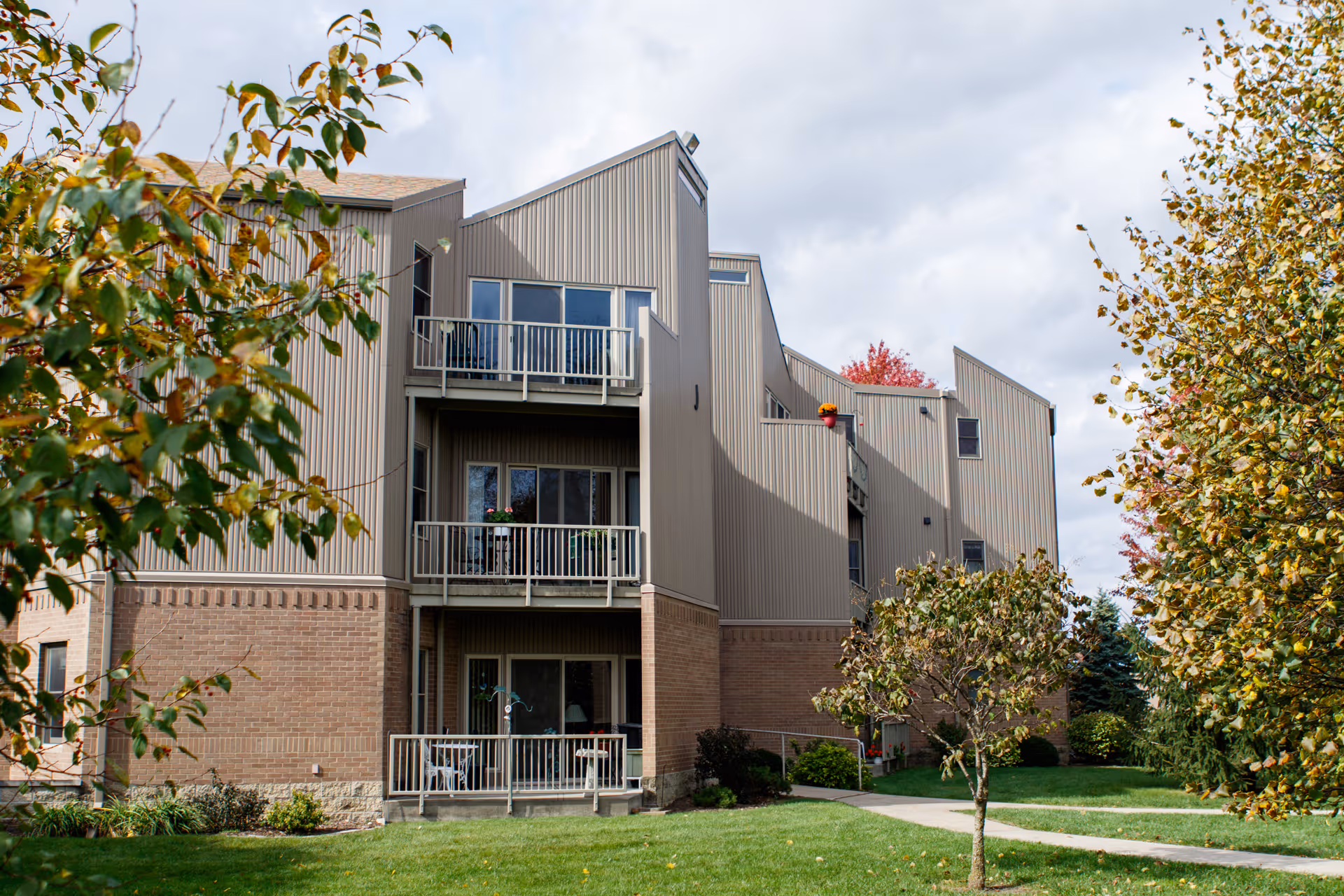 Exterior view of a multi-story residential building with balconies, surrounded by green grass, trees with autumn leaves, and a paved walkway leading to the entrance.