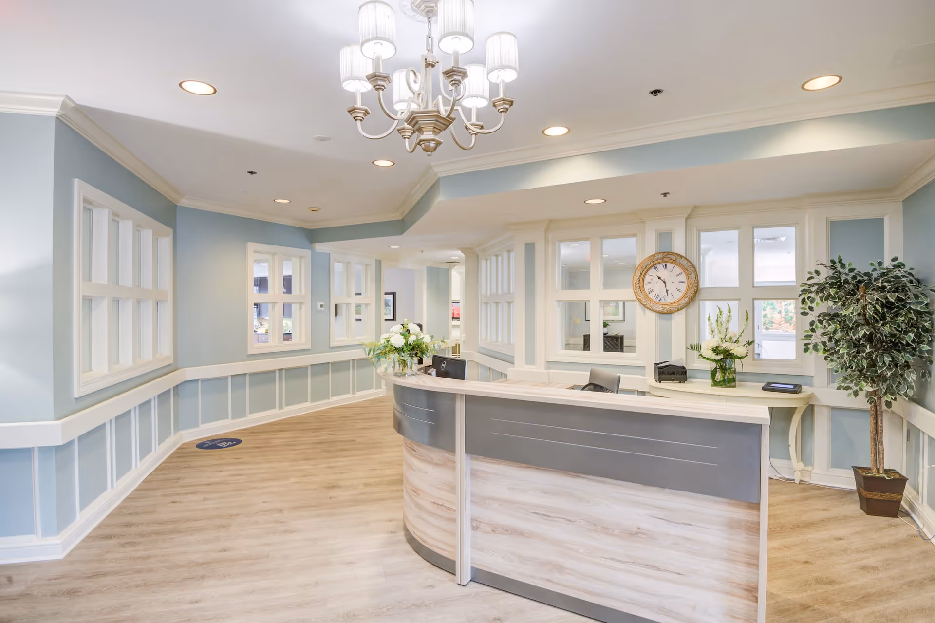 A bright and welcoming reception area in a senior living facility with a curved front desk, light wood flooring, soft blue walls with white trim, a chandelier hanging from the ceiling, a large clock on the wall, and decorative plants and flowers on the desk and in the corner.
