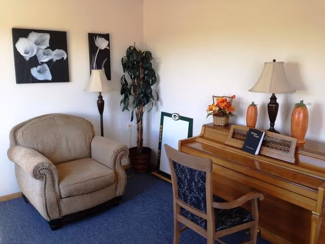 A cozy room with a beige upholstered armchair, a wooden piano with a chair in front of it, two table lamps with beige shades, decorative orange vases, a basket with artificial flowers, a tall potted plant, and two black and white floral paintings on the wall.