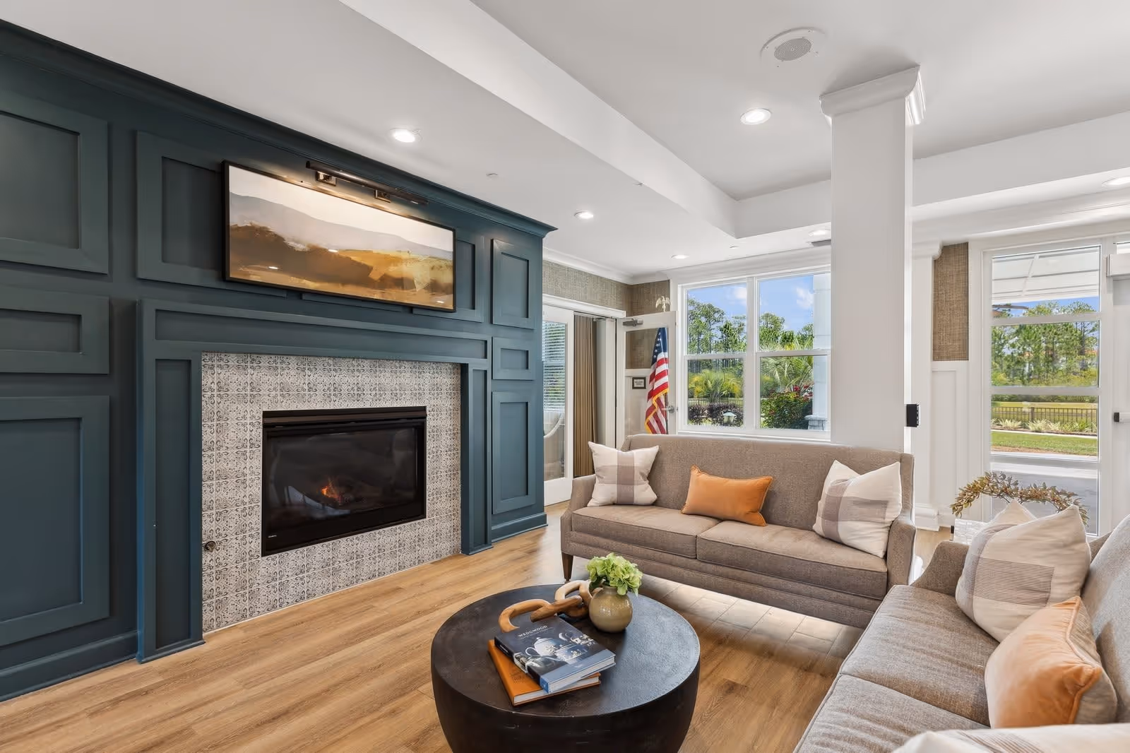 A cozy living room with two beige sofas adorned with pillows, a round black coffee table with books and a small plant, a modern fireplace with a patterned tile surround, and a large window letting in natural light. An American flag is visible near the door, and the room features wood flooring and recessed ceiling lights.