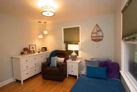 A cozy room with a brown recliner chair next to a white nightstand with a lamp. There is a white dresser with framed photos and decorative items on top. A blue daybed with multiple pillows is positioned under a window with closed blinds. The walls are light-colored and there is a wooden sailboat wall decoration.