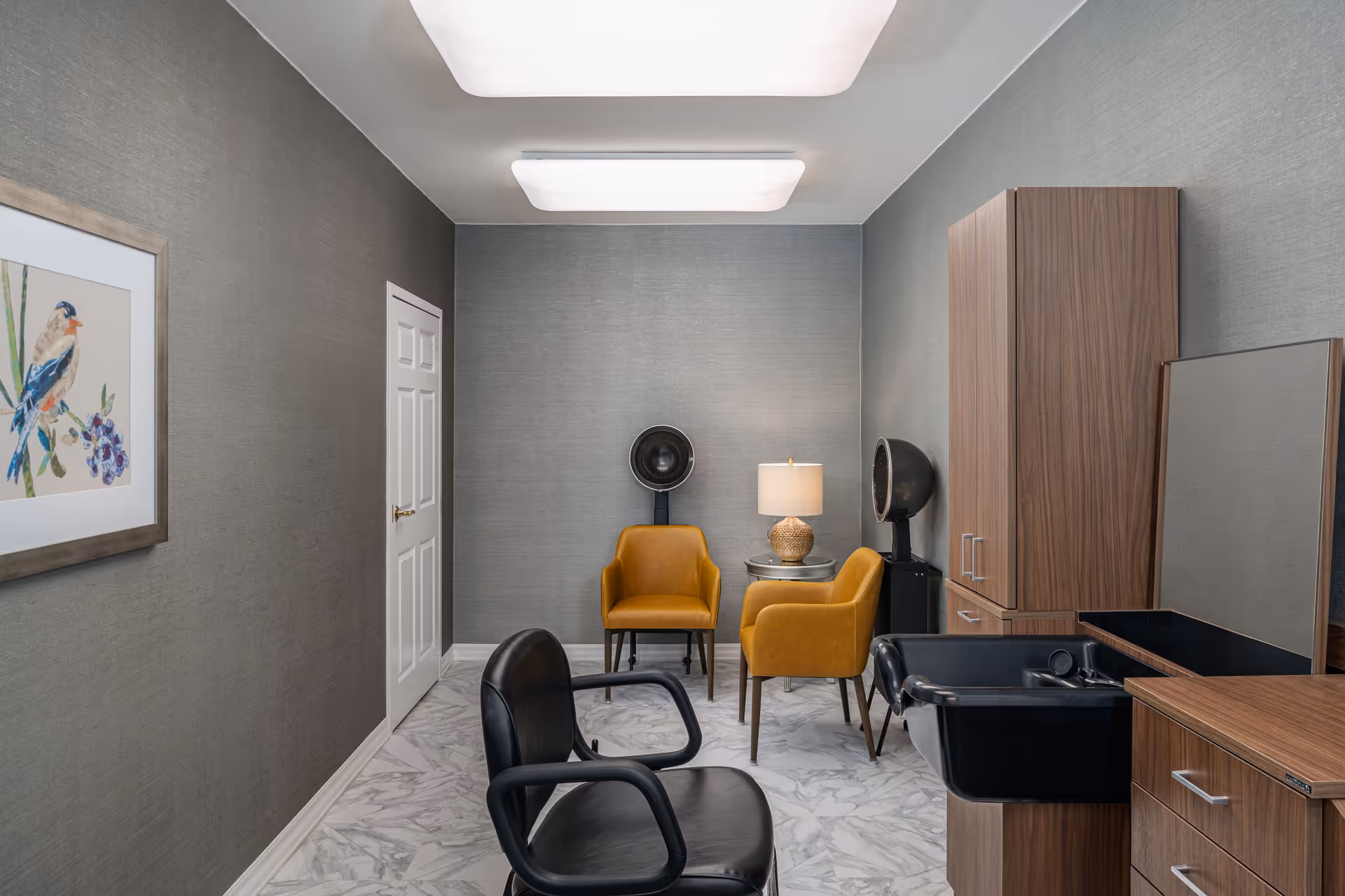 A modern hair salon room with a black salon chair in the foreground, two mustard yellow chairs near the back wall, a small round table with a lamp, two hair dryers, wooden cabinets, and a black sink. The walls are gray with a framed bird artwork on the left wall and the floor has a white and gray marble pattern.