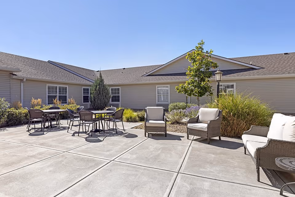 Outdoor patio area at Brookdale Arvada with several wicker chairs and tables arranged on a concrete surface, surrounded by landscaping including bushes, a small tree, and ornamental grasses, under a clear blue sky.