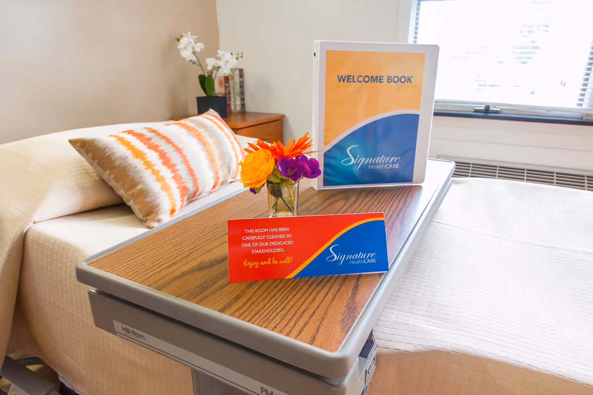 A neatly made bed with a patterned pillow and a bedside table with a small vase of colorful flowers, a welcome book, and a card indicating the room has been carefully cleaned. The room has a window with blinds and a heating unit beneath it.