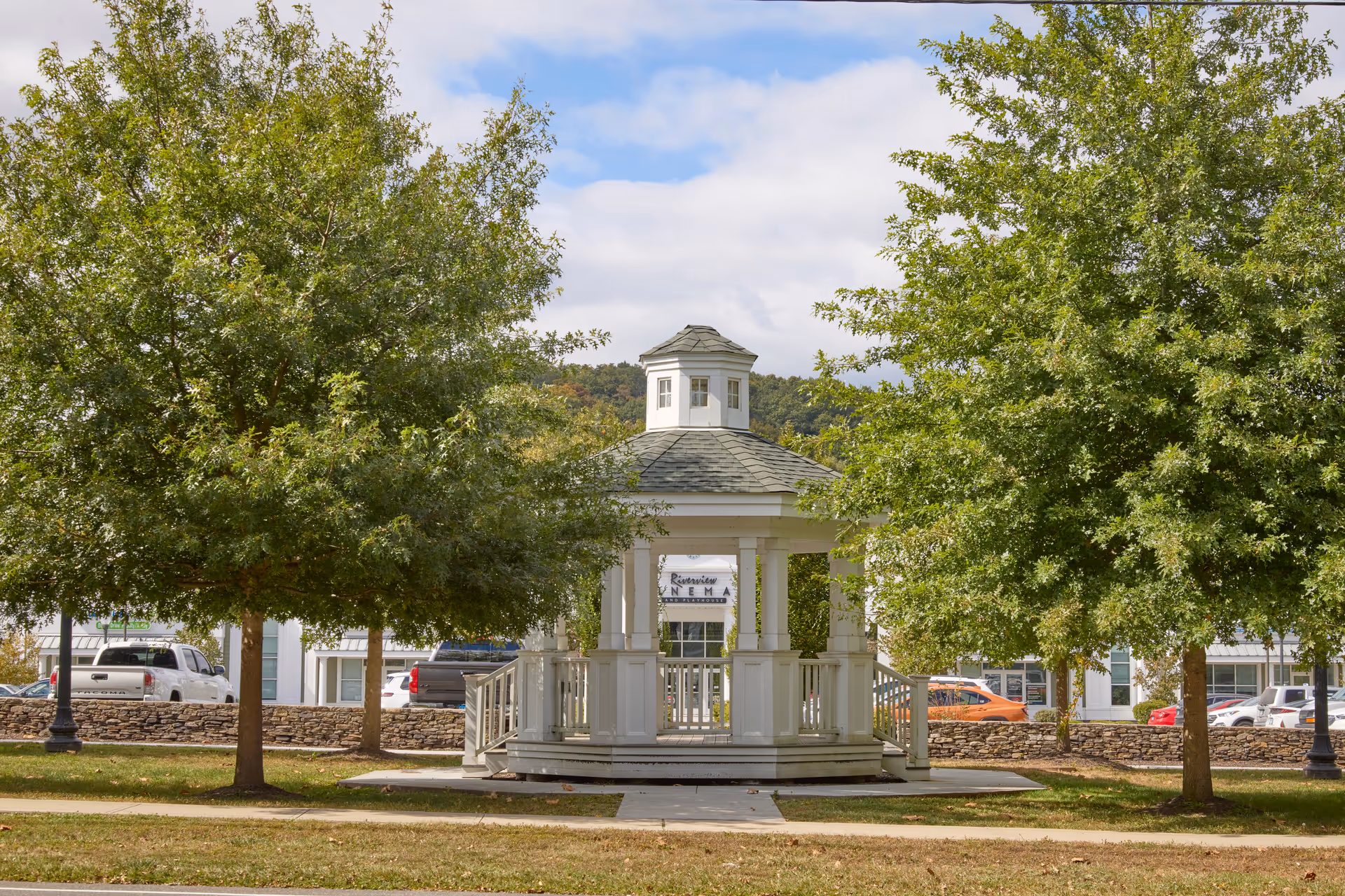 A white gazebo with a small cupola on top, situated between two leafy green trees in a grassy area. Behind the gazebo, there is a low stone wall and parked cars, with a building partially visible in the background under a partly cloudy sky.