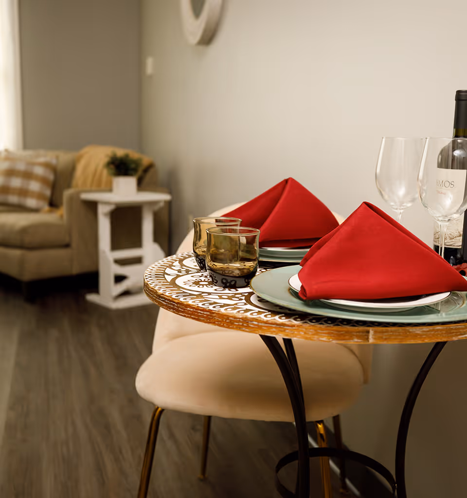 Small bistro table set with plates, red folded napkins, glasses and a wine bottle in a cozy living area.