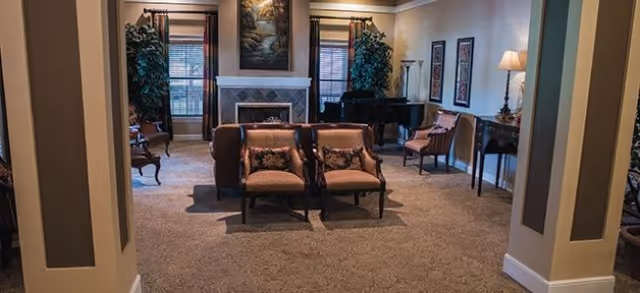 Carpeted communal living room with chairs and sofas arranged facing a fireplace, a piano, lamps, and framed artwork.