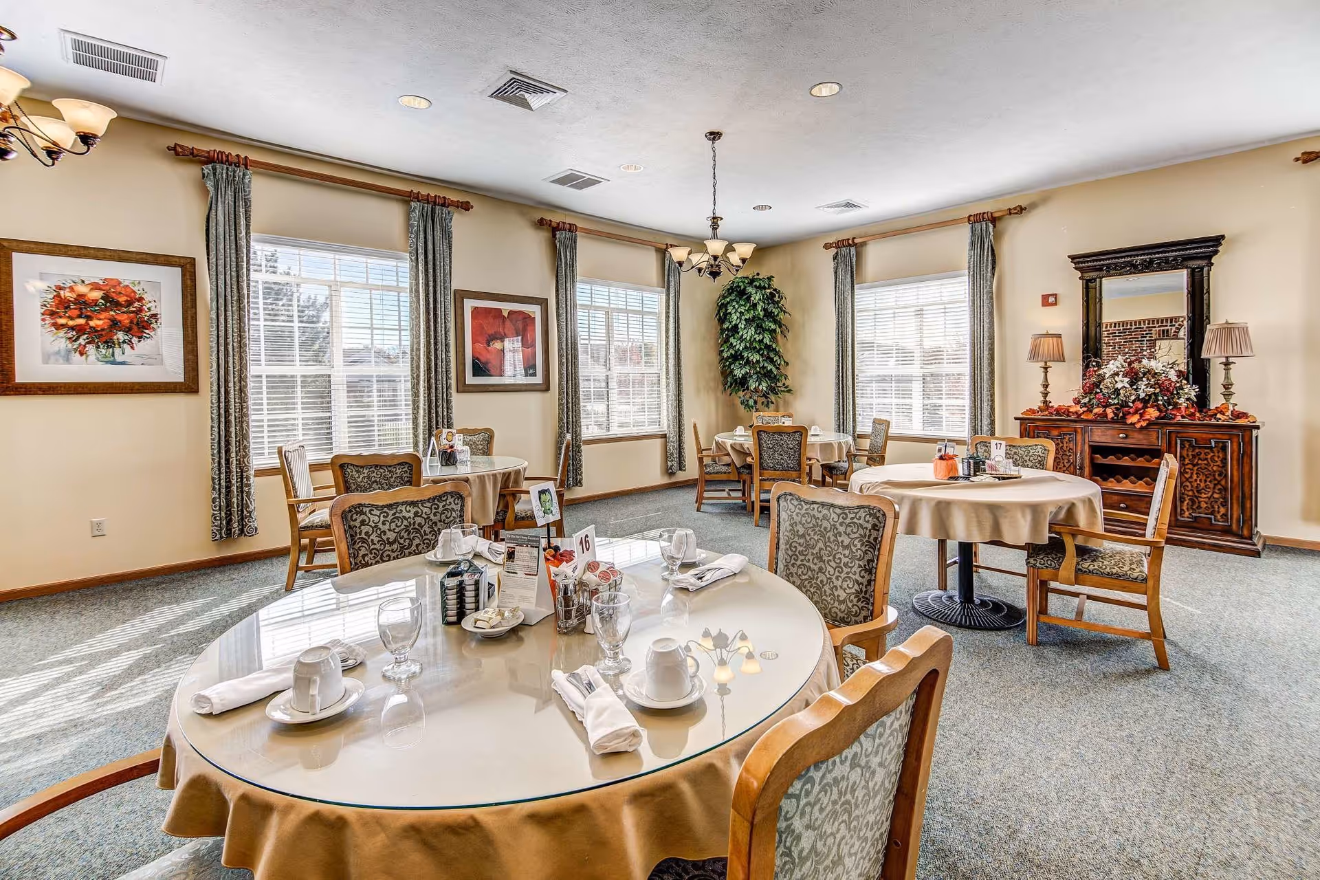 Dining room with round tables set for place settings, upholstered chairs, large windows, and decorative furnishings.