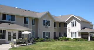 Exterior view of a two-story senior living facility building with beige siding, multiple windows, and a well-maintained lawn with shrubs. There is a patio area with outdoor furniture and an umbrella on the left side.