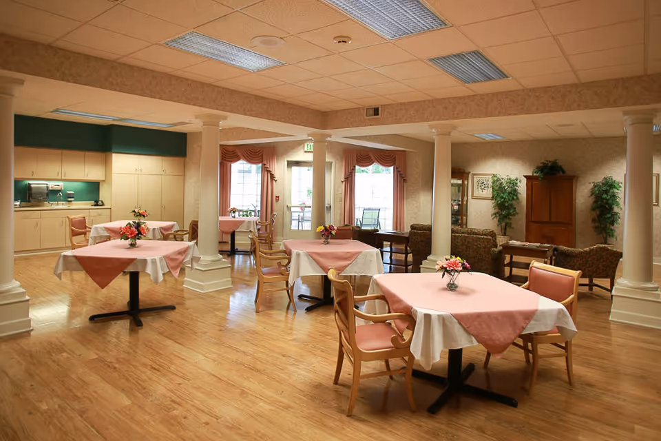 A dining area with several square tables covered with white and pink tablecloths, each adorned with a small flower arrangement. Wooden chairs with pink cushions surround the tables. The room features wooden flooring, white columns, and a kitchenette area with cabinets and a sink in the background. Large windows with pink curtains allow natural light to enter, and there is a seating area with armchairs and a wooden cabinet in the back corner.