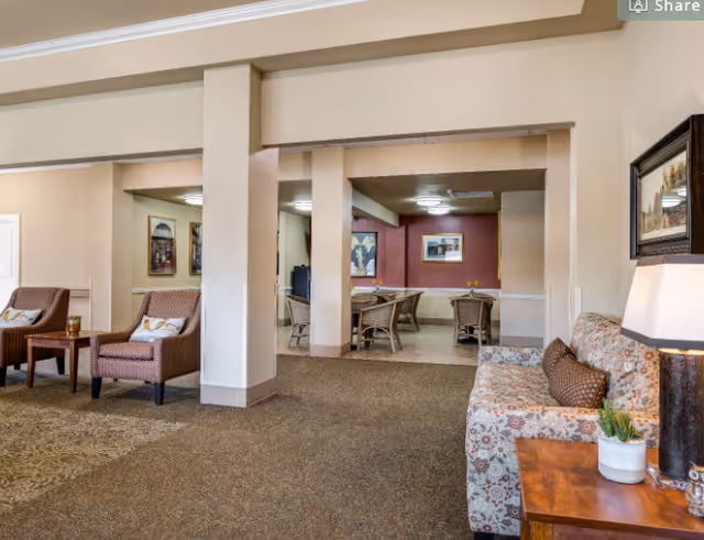Interior view of a senior living facility common area with a patterned sofa and wooden side table with a lamp and plant on the right. Two cushioned armchairs with a small table between them are on the left. In the background, there is a dining area with wicker chairs and tables, and framed artwork on the walls.