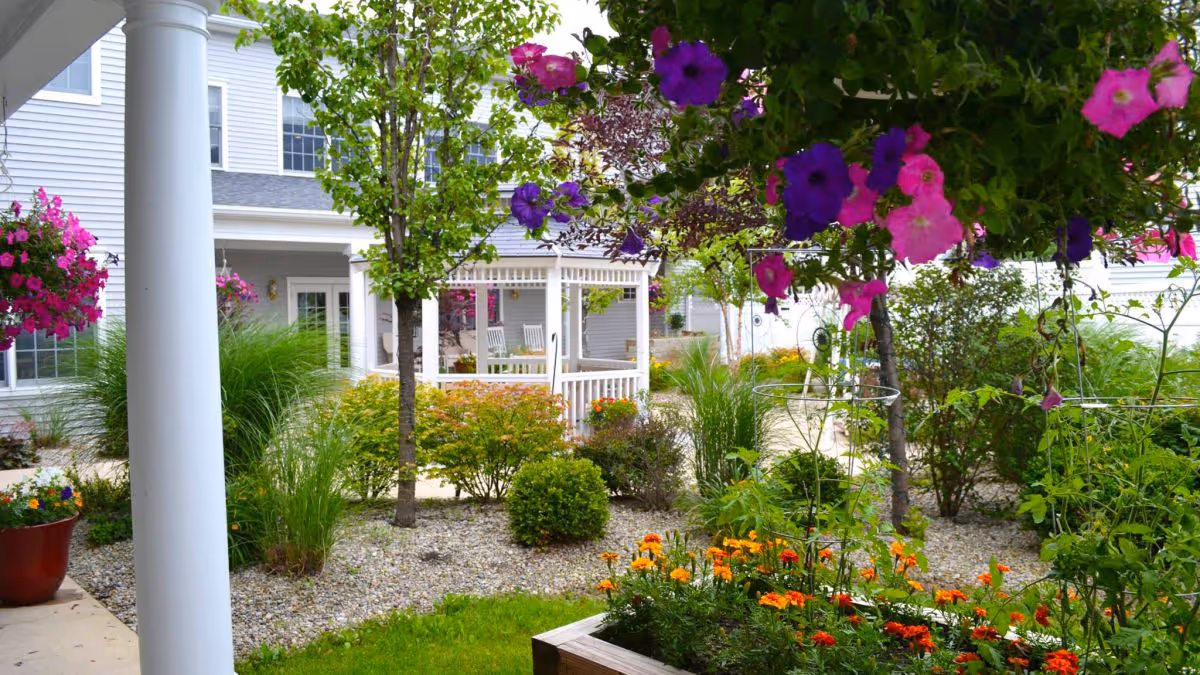 A vibrant outdoor garden area at Kingston Residence of Sylvania featuring colorful flowers, green shrubs, small trees, and a white gazebo with rocking chairs in the background. The scene is bright and inviting with a well-maintained landscape.