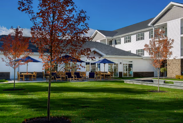 Outdoor patio area with tables, chairs, and blue umbrellas surrounded by a well-maintained lawn and young trees with autumn-colored leaves, adjacent to a multi-story white building under a clear blue sky.