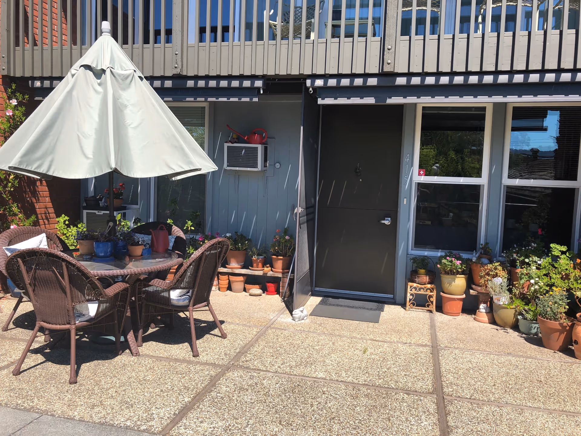 Outdoor patio area with a round glass table surrounded by four wicker chairs with cushions. A large white patio umbrella is positioned over the table. The patio is decorated with numerous potted plants along the wall and near the entrance door of a building. The door is dark-colored with the number 415 displayed vertically beside it. There is an air conditioning unit mounted on the wall above the plants.