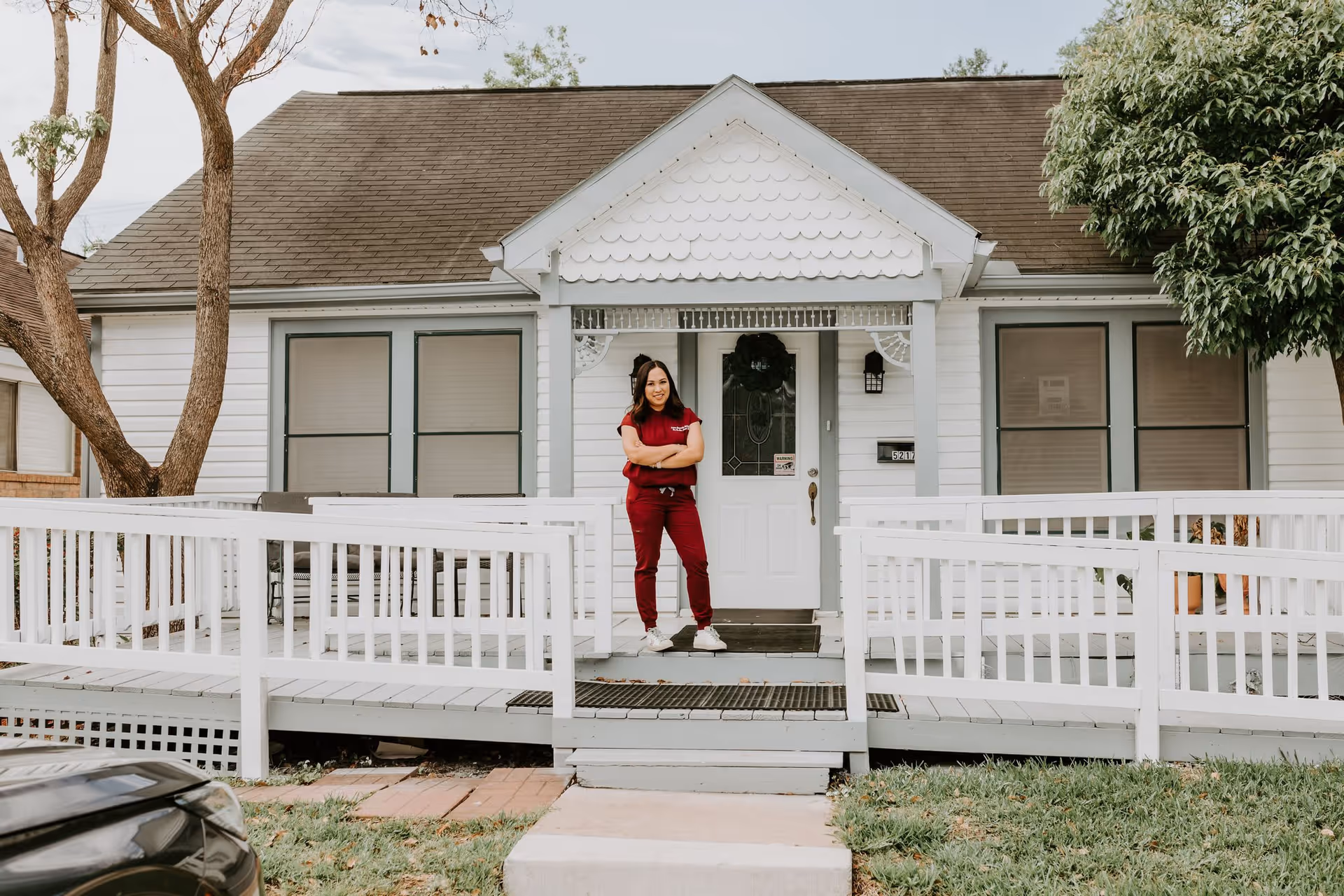 A woman in a red uniform stands with her arms crossed on the front porch of a white house with a ramp and steps leading up to the door. The house has a gabled roof, two windows on either side of the door, and a wreath hanging on the door. There are trees on both sides of the house and a car partially visible in the foreground.