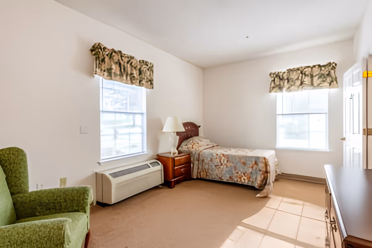 Sunlit bedroom with a single bed, nightstand and lamp, a green armchair, and two windows.
