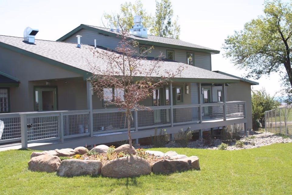 Exterior view of a single-story assisted living facility building with a sloped roof and a ramped porch railing. In the foreground, there is a small landscaped area with rocks and a young tree surrounded by grass. Trees and a fence are visible in the background under a clear sky.