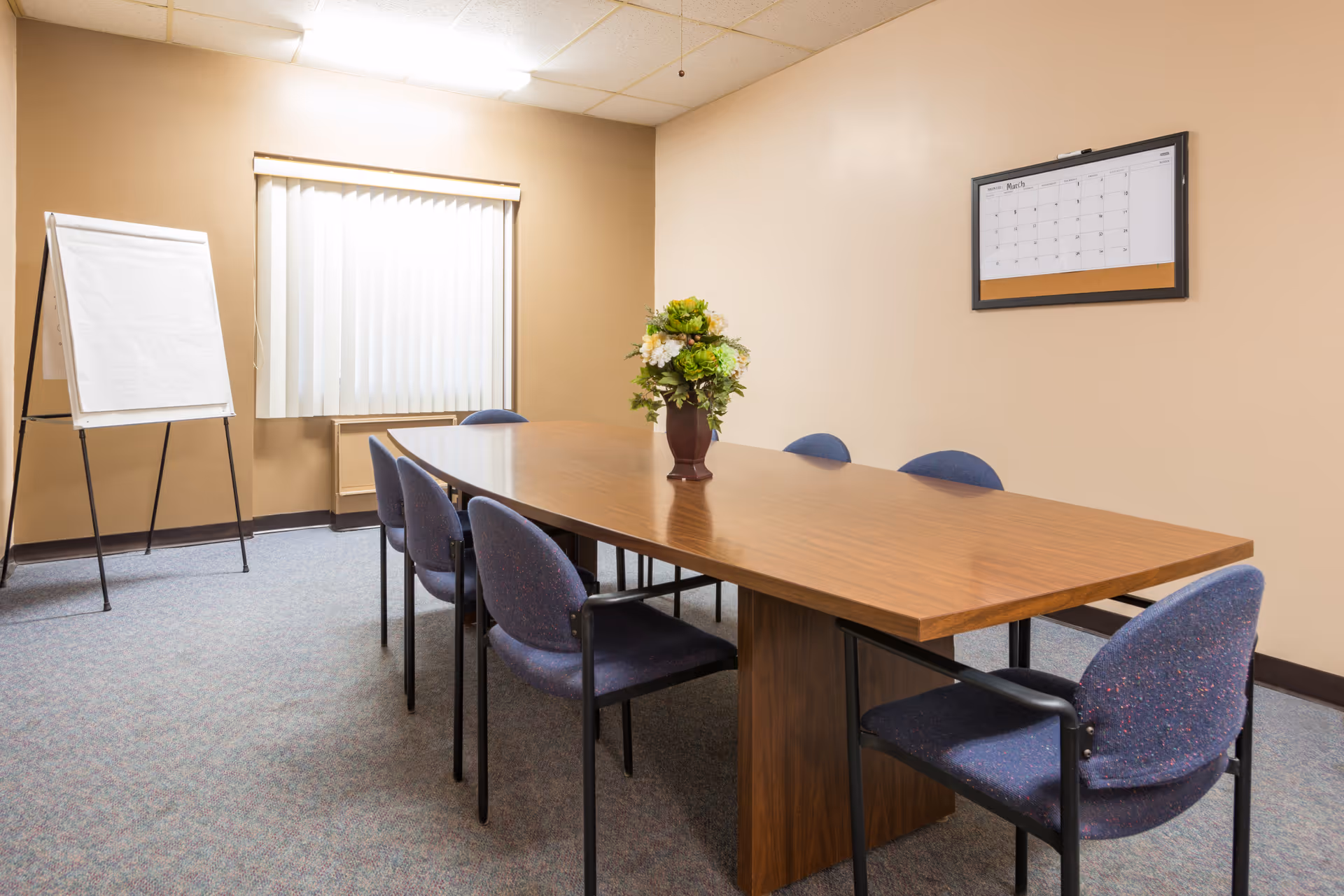 A conference room with a long wooden table surrounded by six blue upholstered chairs. A vase with green and white flowers is placed in the center of the table. On the left side of the room, there is a window with vertical blinds and a flip chart stand. A wall calendar is mounted on the beige wall to the right.